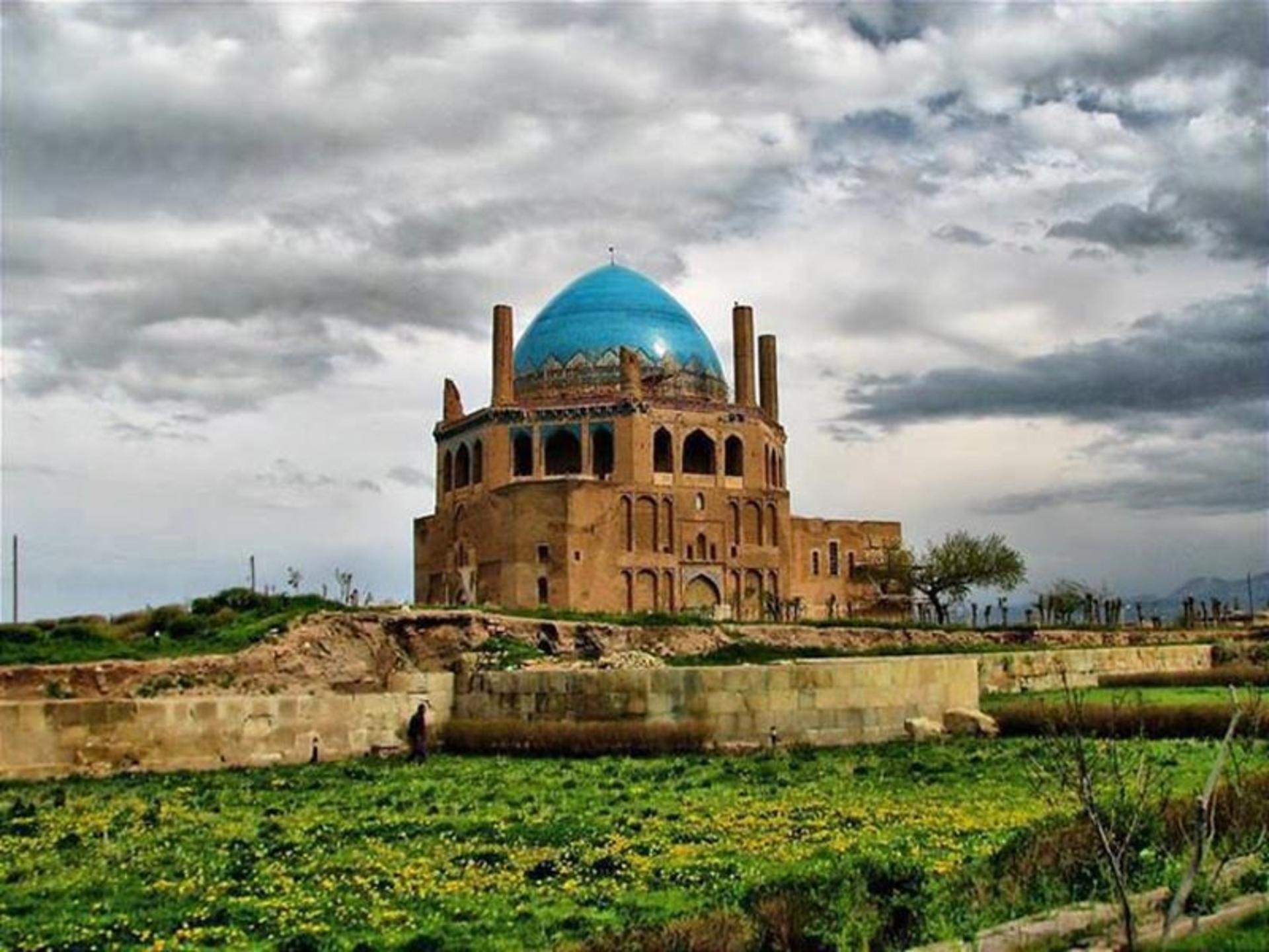 نمایی از بیرون گنبد سلطاننیه در میان دشت سبز و آسمان پر ابر