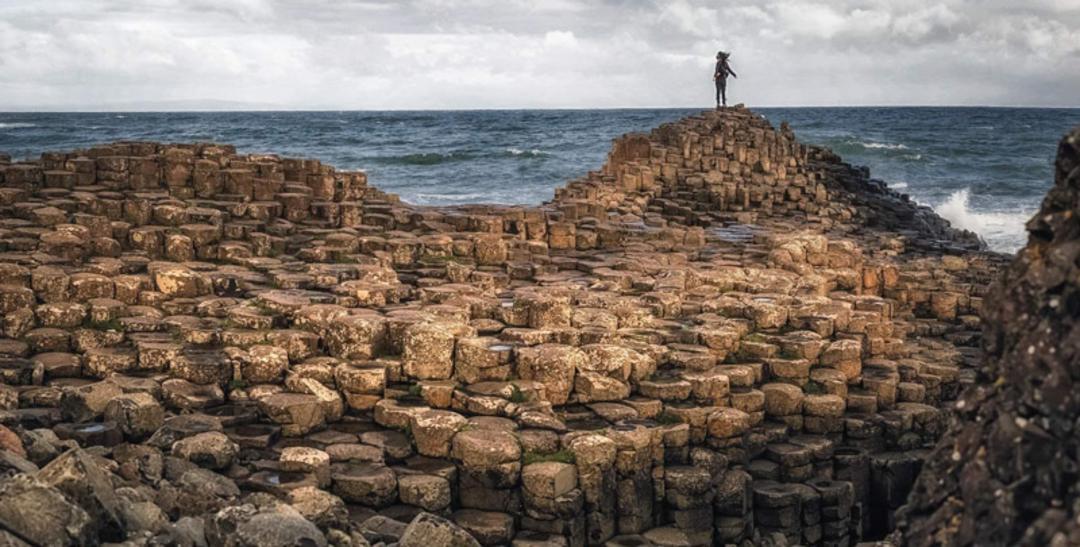 فردی ایستاده بر سنگ های استوانه ای گذرگاه ساحلی جاینت؛ منبع عکس: Ireland.com، عکاس: نامشخص