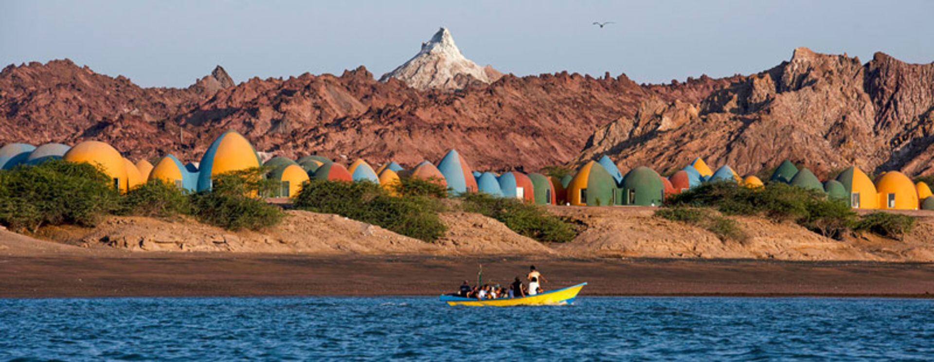 قایق سواران در جزیره هرمز با پس زمینه اقامتگاه رنگارنگ ماجرا؛ منبع عکس: Fair Tourism، عکاس: Tahmineh Monzavi