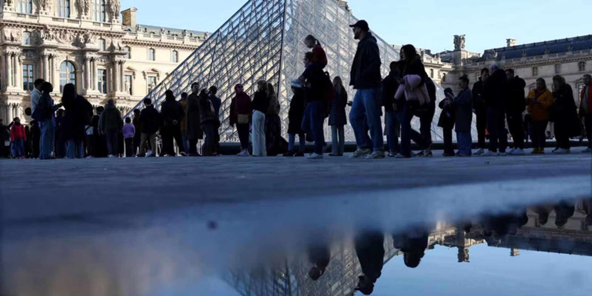 صف مردم جلوی هرم لوور، بازتاب روی زمین مرطوب (زاویه پایین)