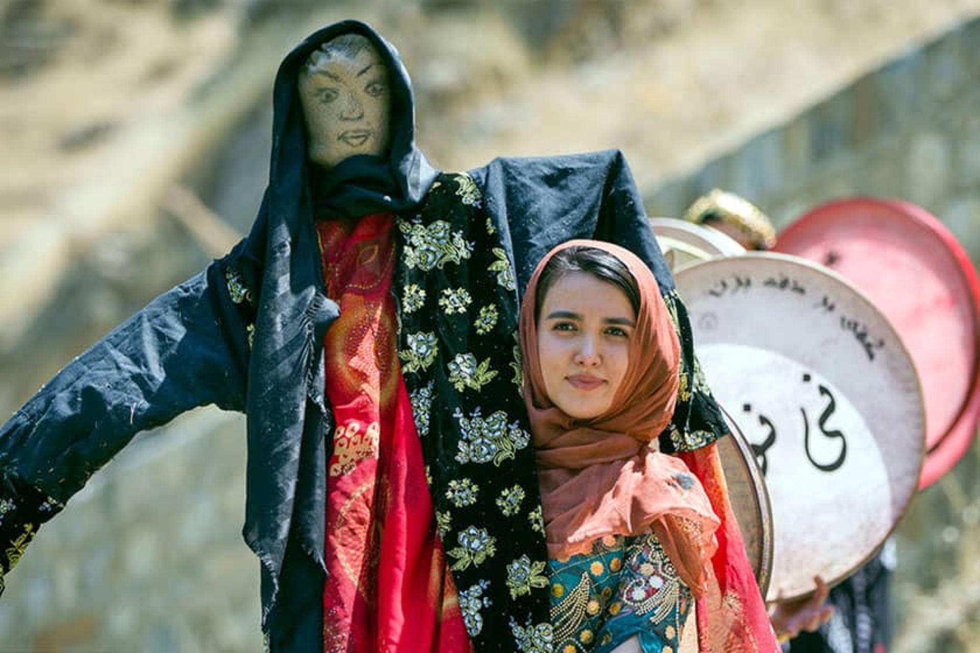دختر جوانی با مترسکی در دست و افرادی پشت سر او در حال زدن دف