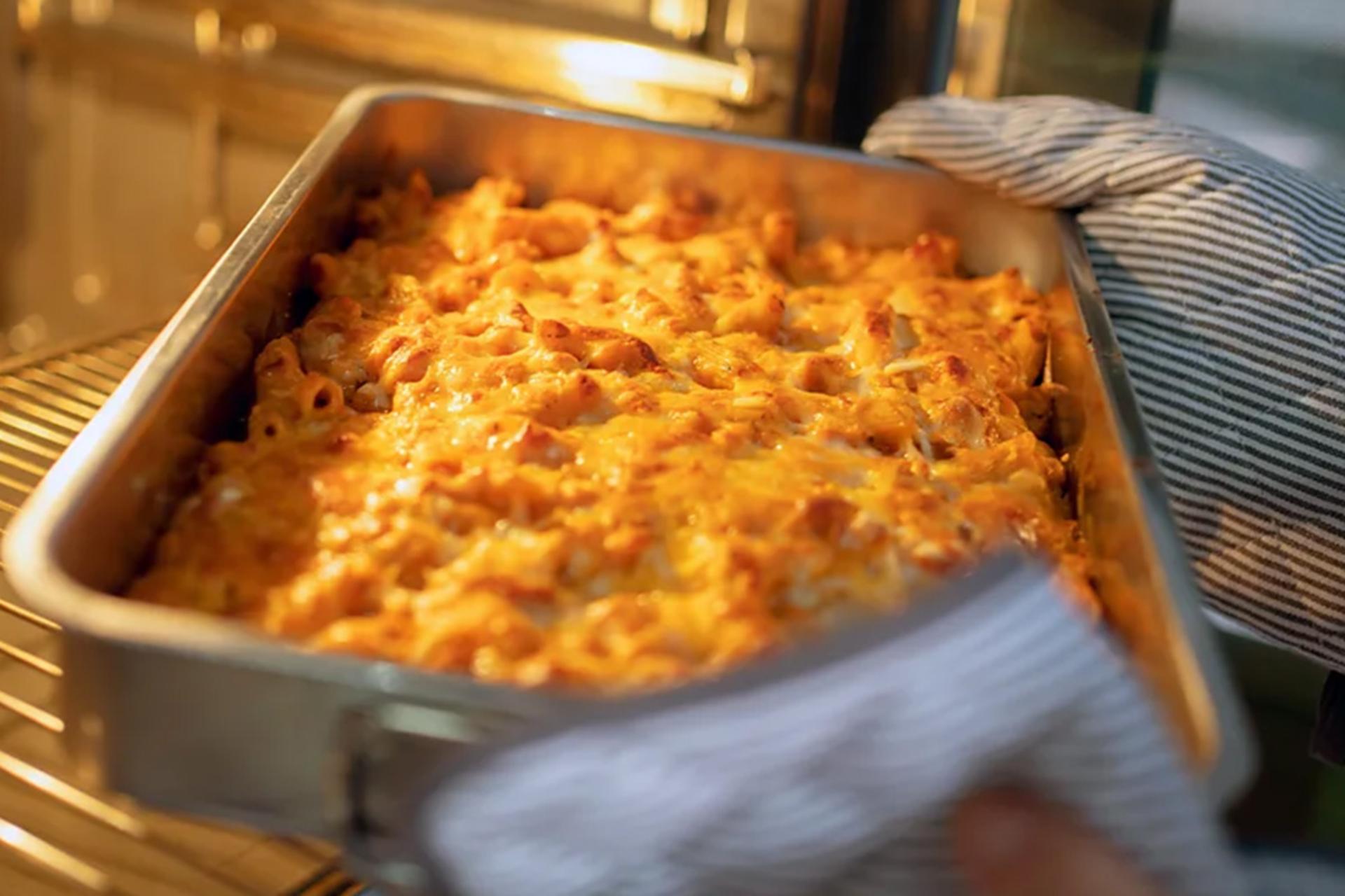 دست‌هایی با دستکش فر در حال بیرون آوردن سینی ماکارونی و پنیر برشته از داخل فر