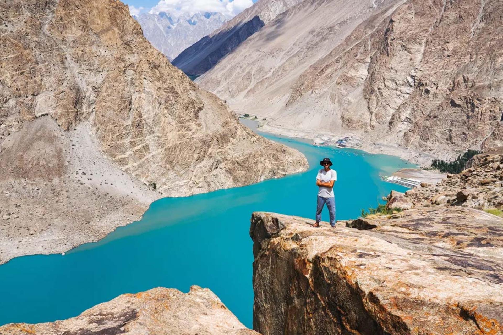 فردی در لبه صخره با نمای دریاچه عطاباد در میان کوه‌های سنگی