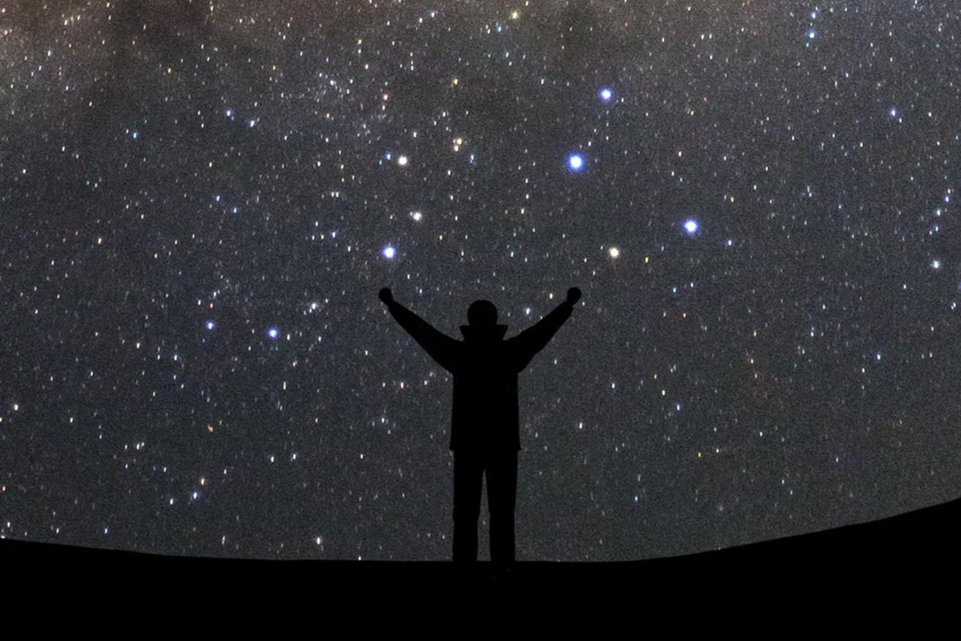 تصویر ضد‌نور از فردی با دستان باز که رو به کهکشان راه شیری ایستاده