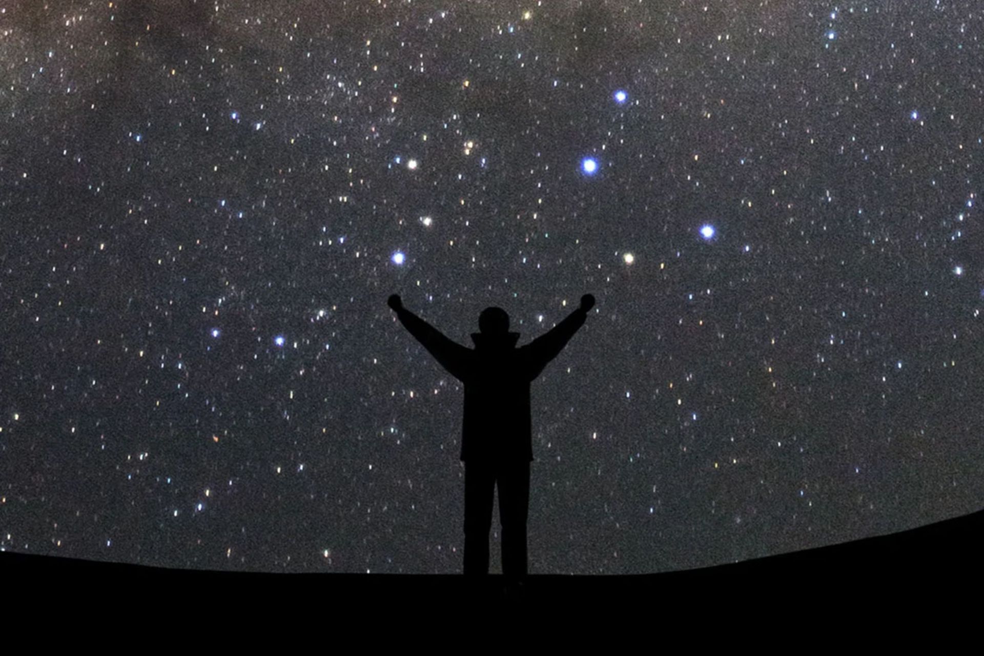 تصویر ضد‌نور از فردی با دستان باز که رو به کهکشان راه شیری ایستاده