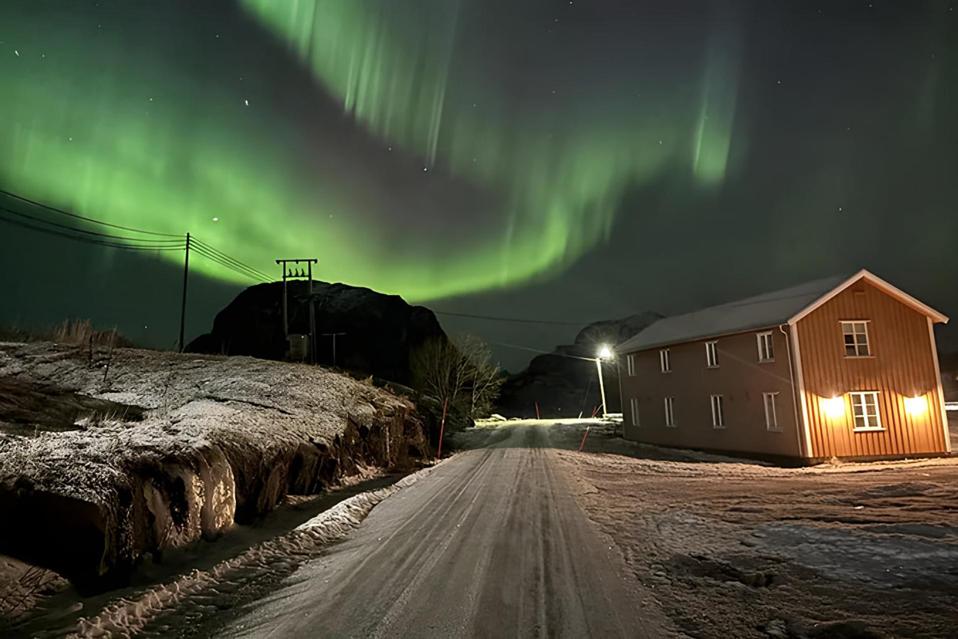 شفق قطبی (Aurora Borealis) سبز رنگ در آسمان شب بالای یک خانه روستایی برفی