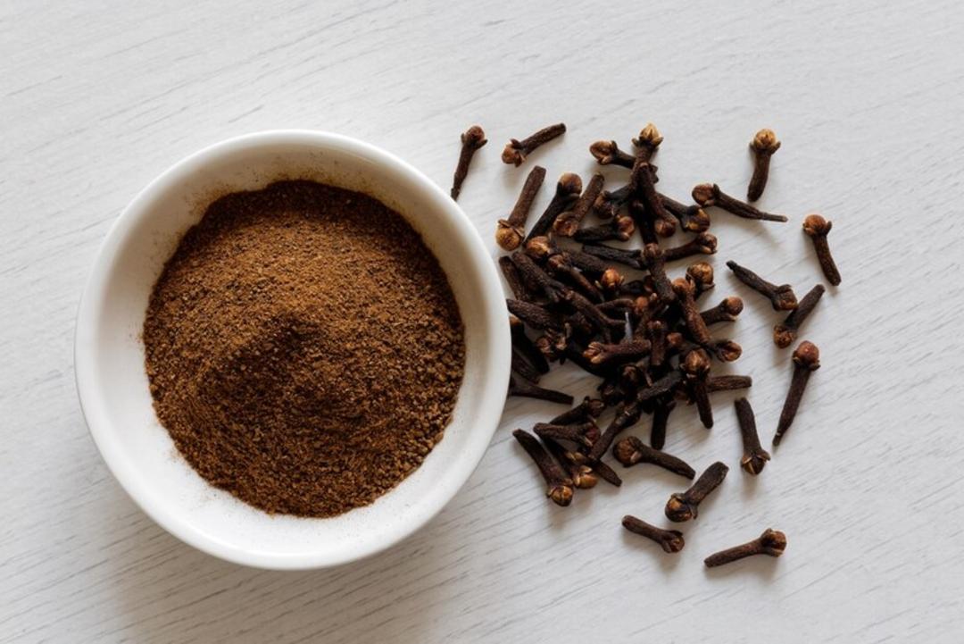 میخک (Clove) پودر شده در ظرف سفید و دانه کامل در کنار آن