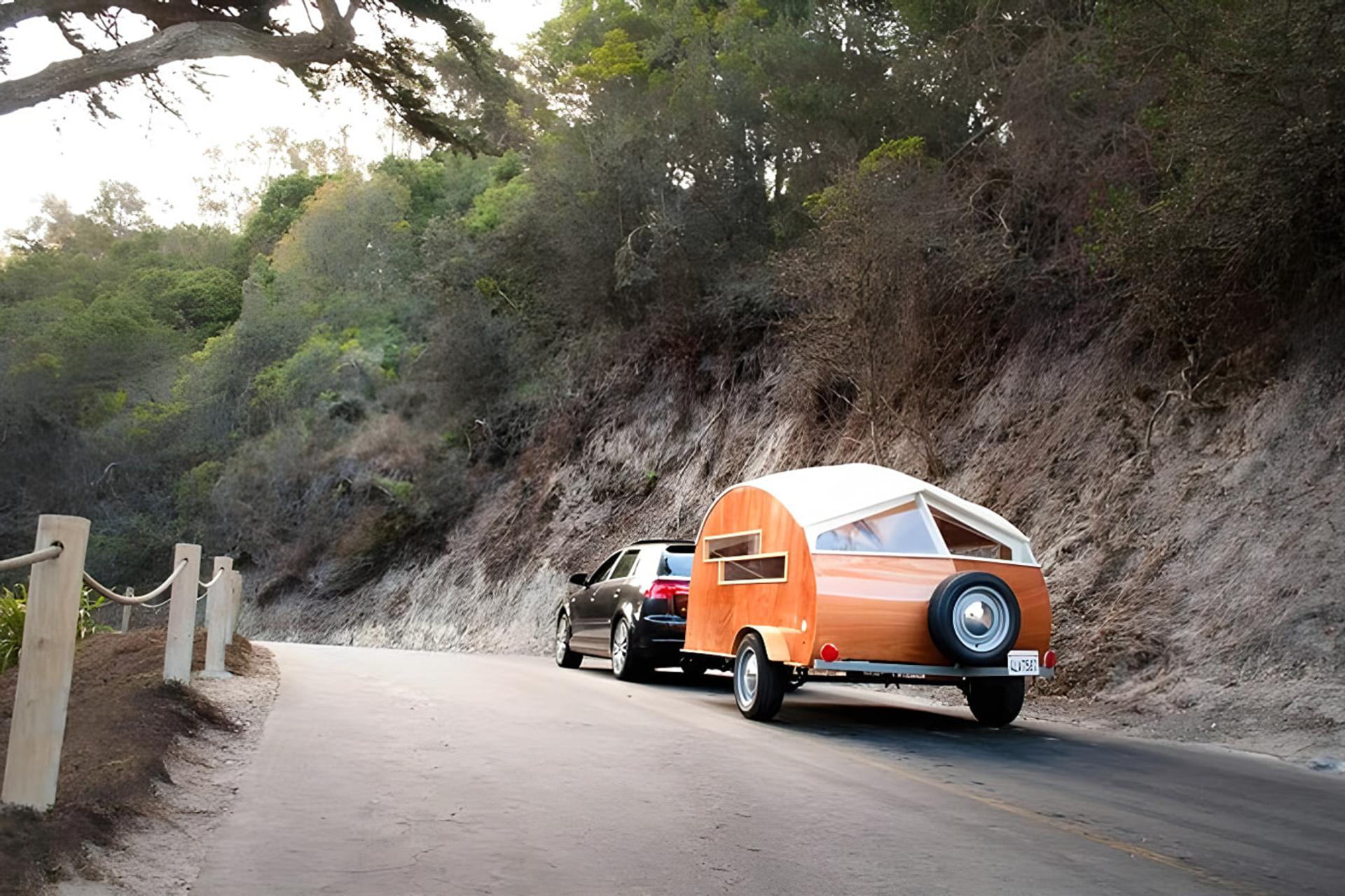 کاروان چوبی (Teardrop Camper) کوچک در حال حرکت در جاده‌ای میان درختان