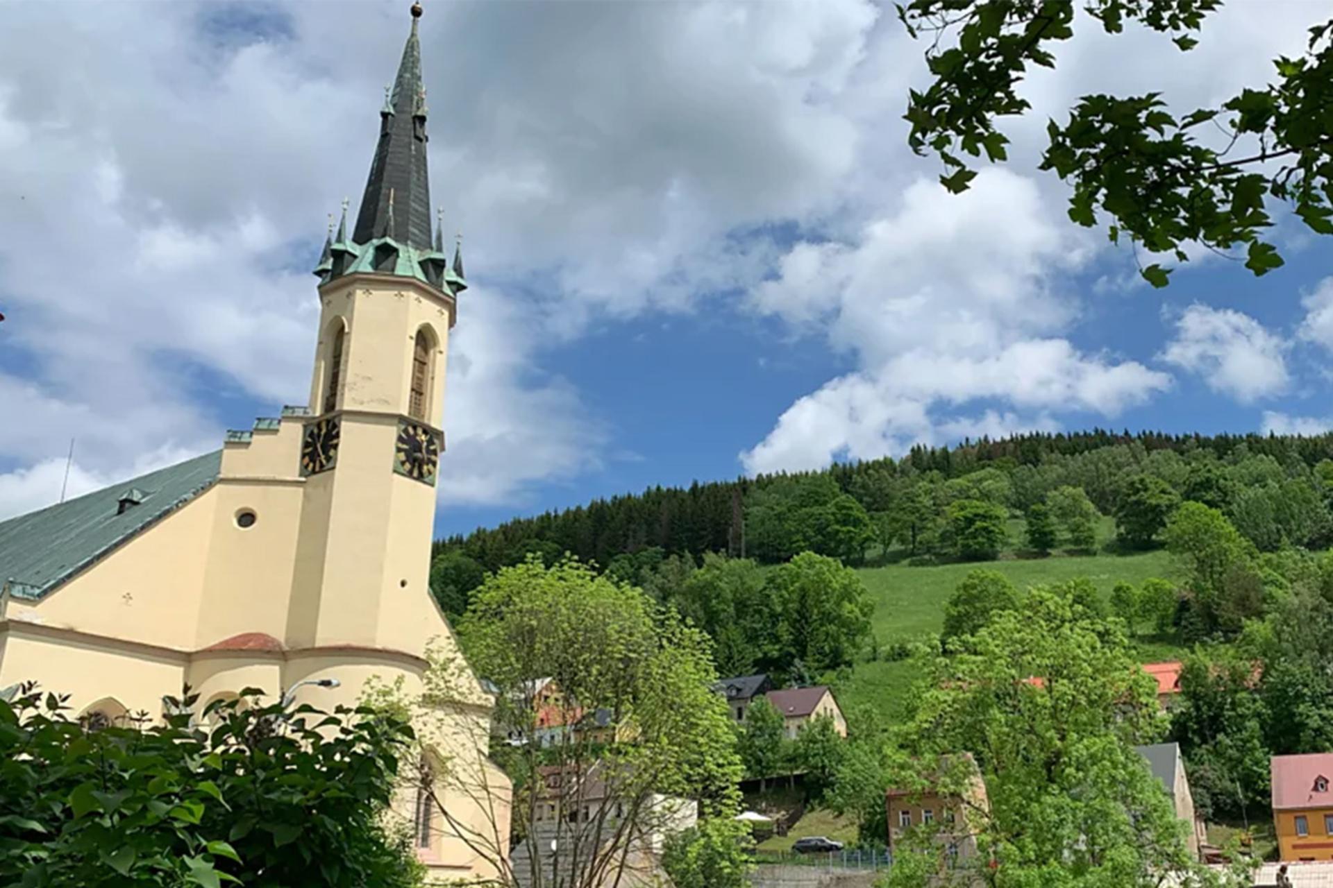 نمای خارجی کلیسای باستانی یاخیموف با برج ناقوس بلند و نوک‌تیز، در میان درختان سرسبز و تپه‌های پوشیده از جنگل.