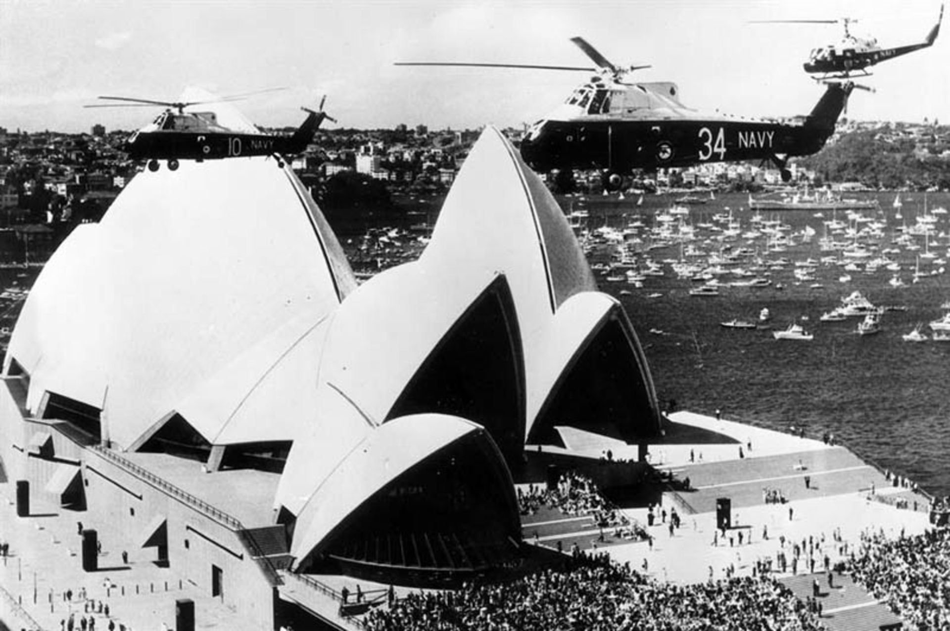 نمای هوایی قدیمی از ساختمان اپرای سیدنی با حضور هلیکوپترهای نیروی دریایی در حال پرواز