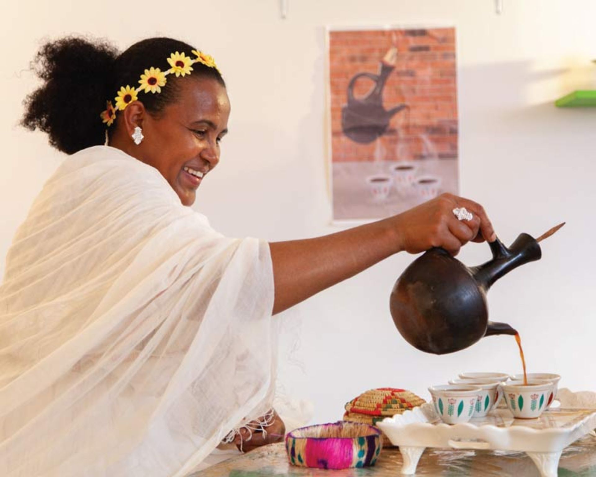 زنی با لباس سفید و سربند گل آفتابگردان در حال ریختن قهوه سنتی اتیوپی
