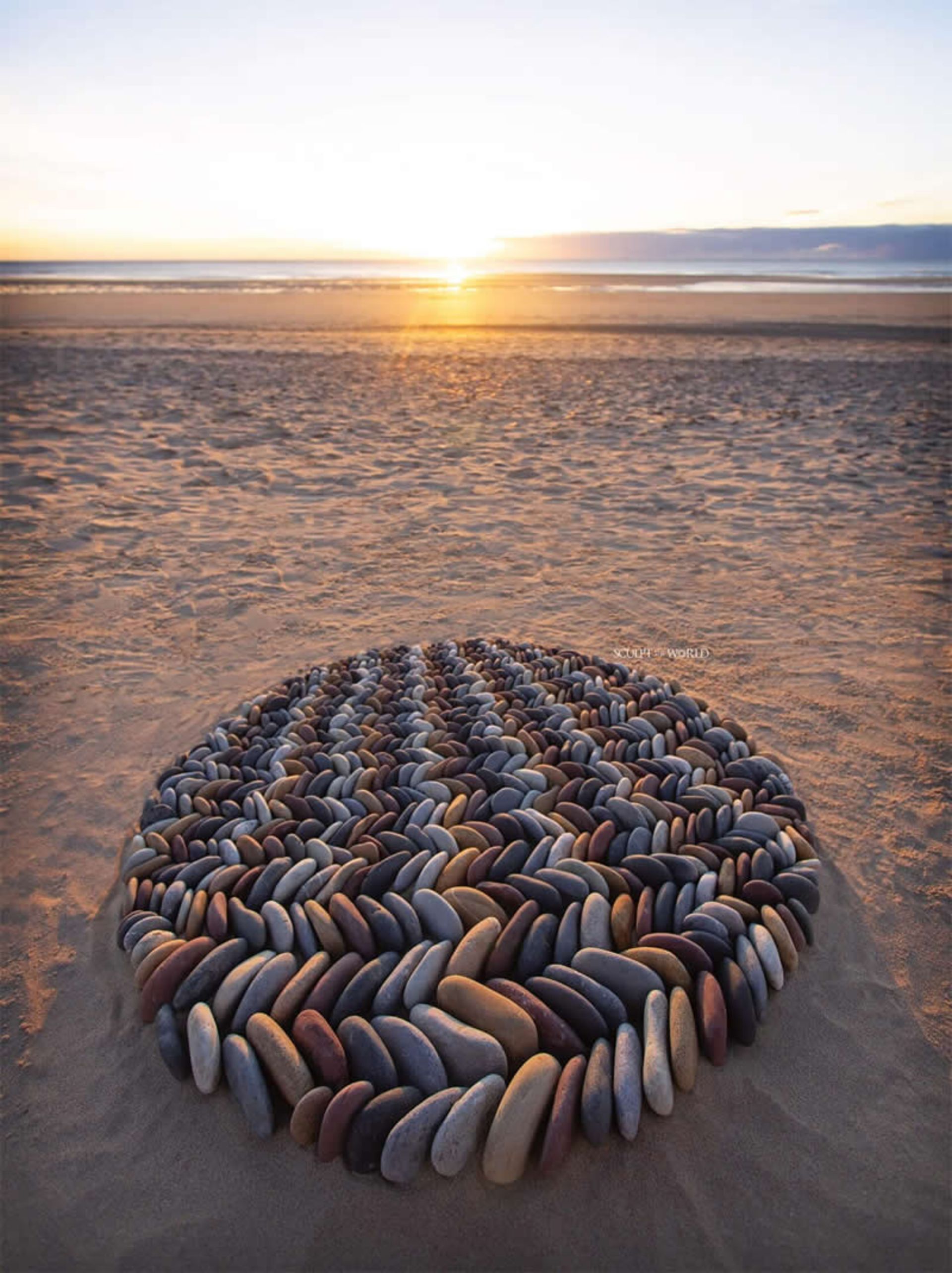 طرح متقارن و دایره‌ای از سنگ‌های روشن و تیره در غروب ساحل