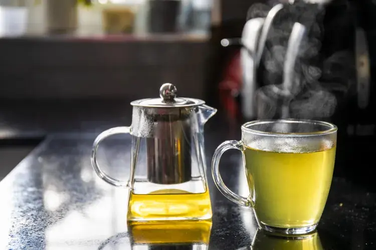 نوشیدنی گیاهی زرد رنگ در فنجان و قوری شیشه‌ای روی میز