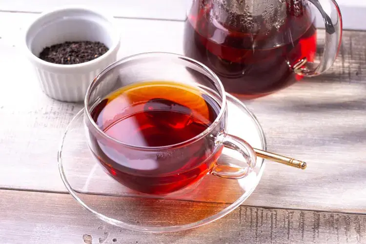 فنجان چای سیاه در کنار قوری و ظرف کوچک برگ‌های چای