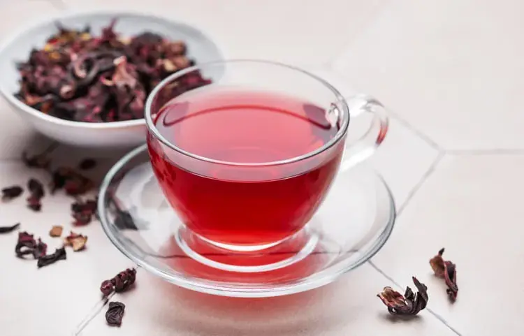چای گل کارکدها (Hibiscus) در فنجان شیشه‌ای در کنار گل‌های خشک