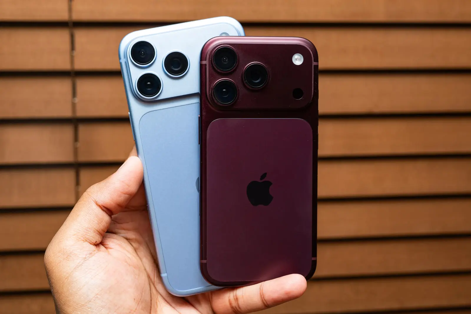 دو گوشی هوشمند آیفون (iPhone) با رنگ‌های آبی و زرشکی در دست یک فرد