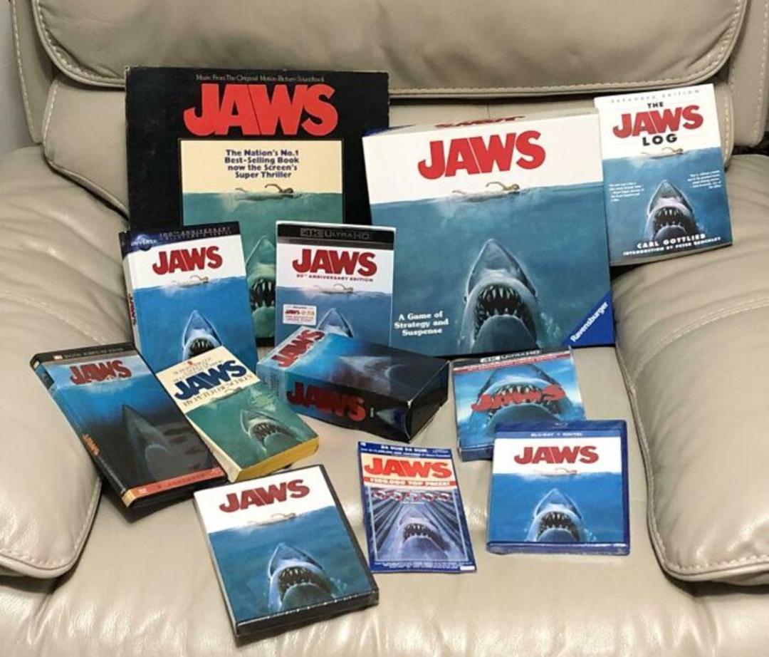 مجموعه‌ای از کتاب‌ها و نسخه‌های مختلف فیلم کوسه (Jaws) روی مبل