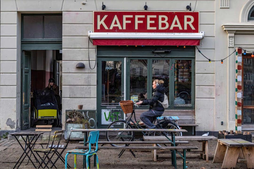 دوچرخه‌سواری در مقابل کافه بار (Kaffebar) با میز و صندلی‌های فضای باز