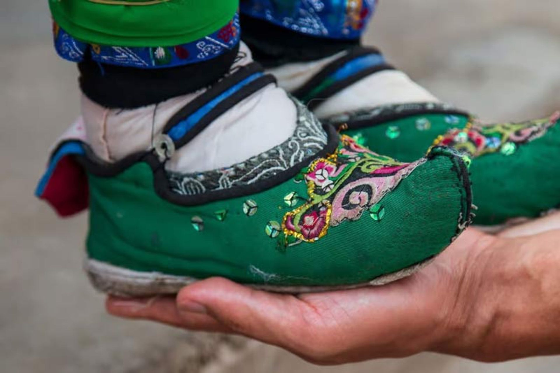 تصویری که تضاد اندازه را نشان می‌دهد؛ دستان یک فرد که یک جفت کفش گلدوزی شده سبز رنگ «لوتوس» را نگه داشته است