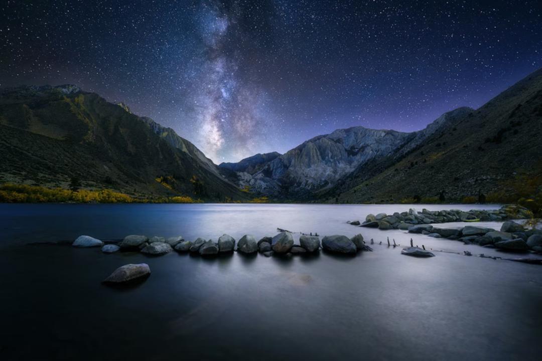 کهکشان راه شیری (Milky Way) در آسمان شب بالای کوه‌های پوشیده از سنگ و دریاچه