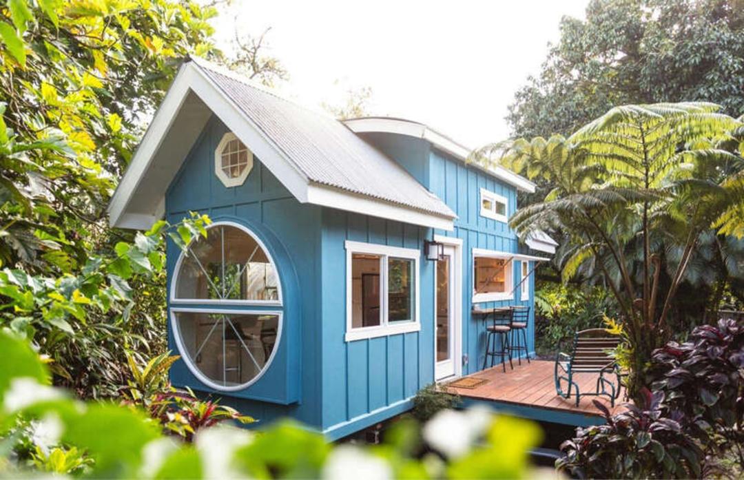 خانه مینیاتوری (Tiny House) آبی رنگ با پنجره‌های بزرگ در میان درختان سبز