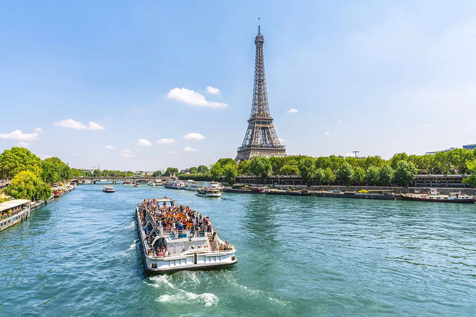 منظره‌ای روشن و آفتابی از رودخانه سن در پاریس با یک کشتی تفریحی پر از گردشگر در حال حرکت روی آن