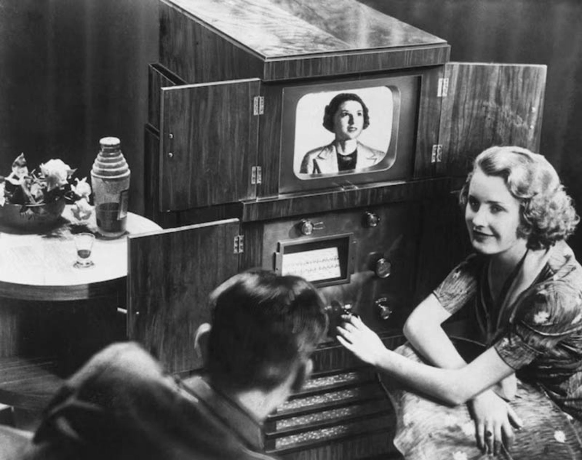 یک دستگاه تلویزیون قدیمی بزرگ و چوبی با درهای بازشو که تصویری از یک زن را نشان می‌دهد
