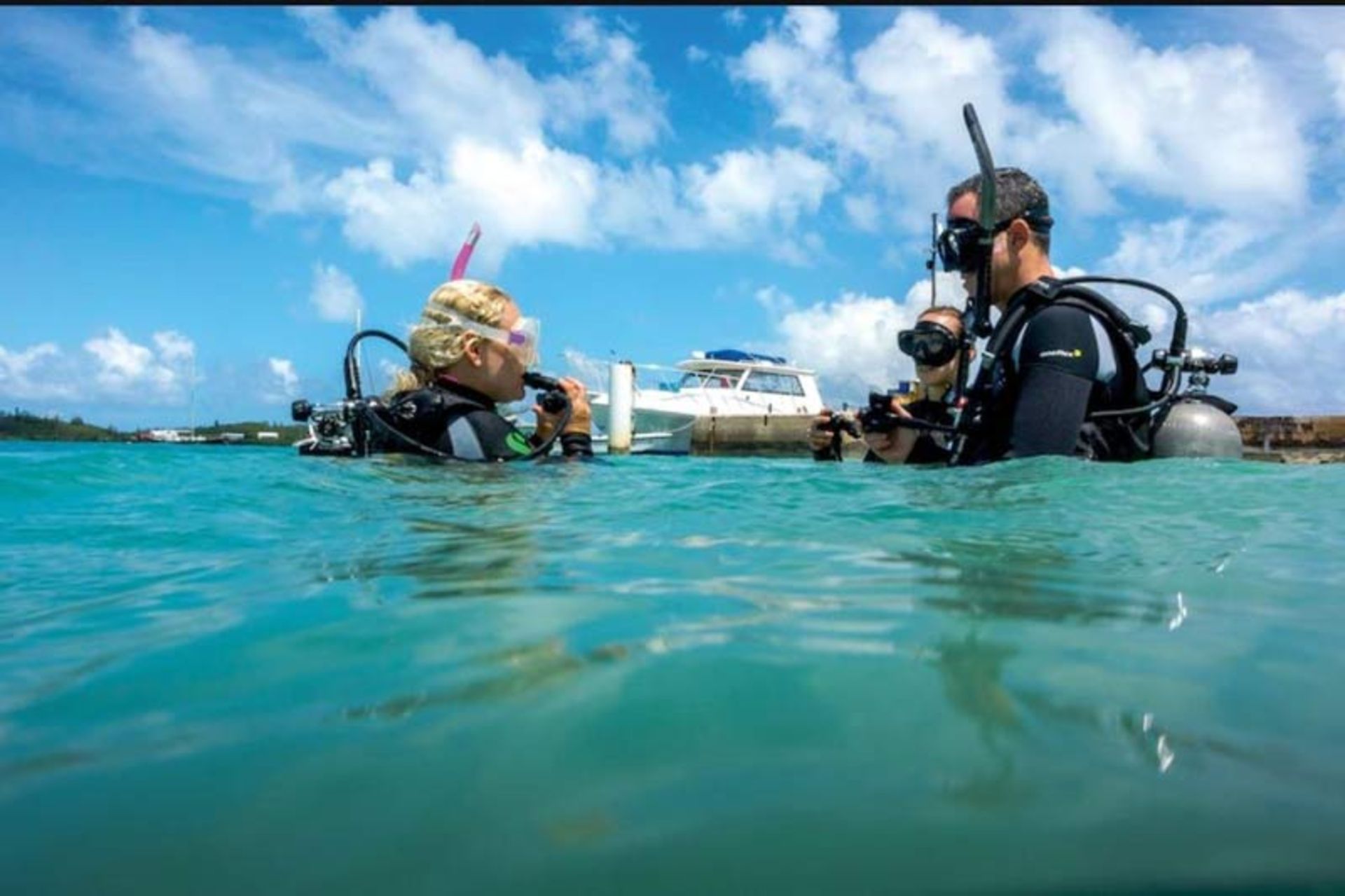غواصان با لباس غواصی و کپسول در حال آماده شدن برای شیرجه در آب‌های کم‌عمق ساحلی در نزدیکی یک قایق.