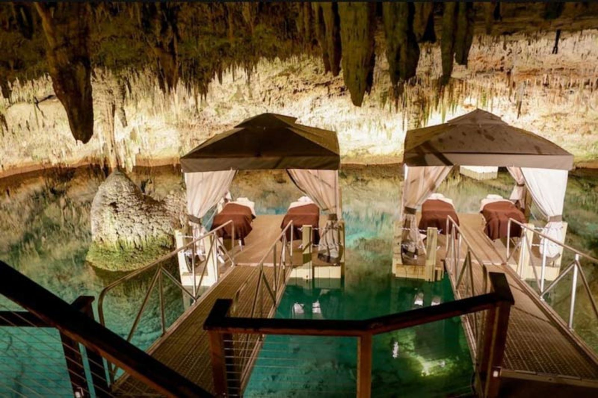 دو تخت ماساژ لوکس با سایه‌بان‌های پارچه‌ای سفید در داخل یک غار ساحلی طبیعی بر فراز آب‌های فیروزه‌ای