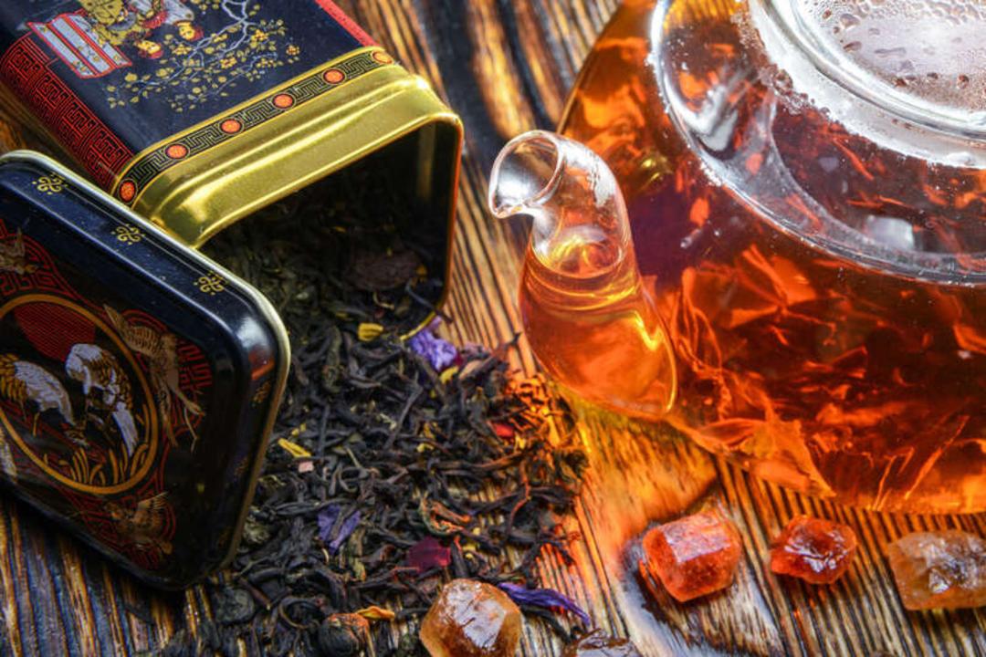 برگ‌های خشک چای در کنار قوری شیشه‌ای و تکه‌های شکر